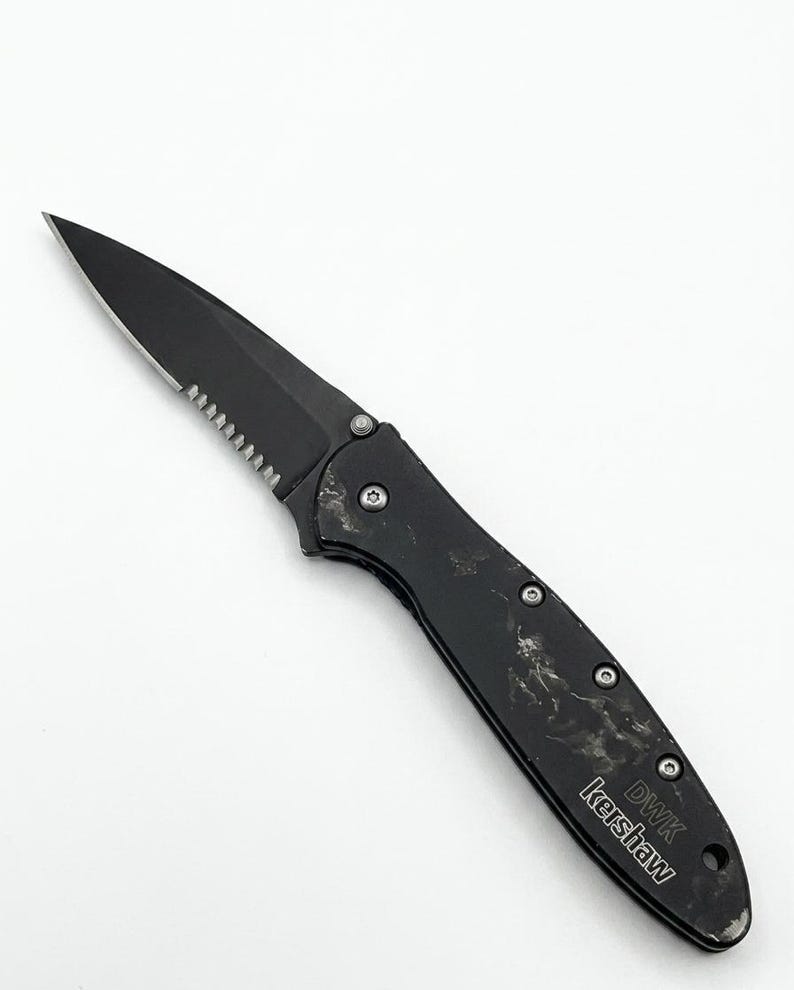 May include: Black folding knife with a serrated edge and a pointed tip. The handle has a textured appearance with visible rivets. The text "DWK Kershaw" is printed on the handle.