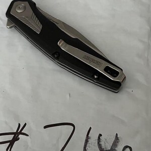 May include: A black and silver folding knife with a pocket clip. The knife has a black handle and a silver blade. The brand name "Kershaw" is visible on the clip. The number "7140" is written in black ink on the white surface.