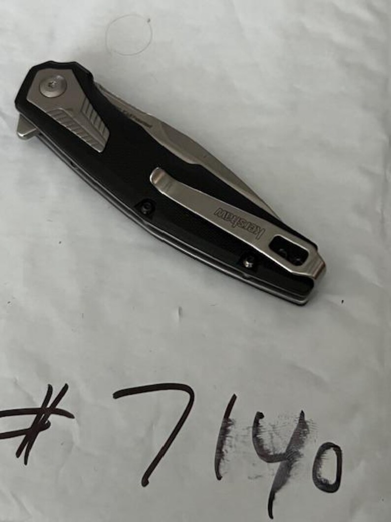 May include: A black and silver folding knife with a pocket clip. The knife has a black handle and a silver blade. The brand name "Kershaw" is visible on the clip. The number "7140" is written in black ink on the white surface.