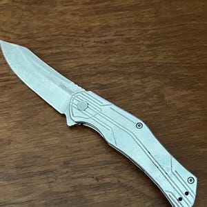 May include: A silver folding knife with a textured blade and handle. The knife has a partially serrated edge and a clip for carrying. The handle features a geometric design and a button for opening and closing the blade.