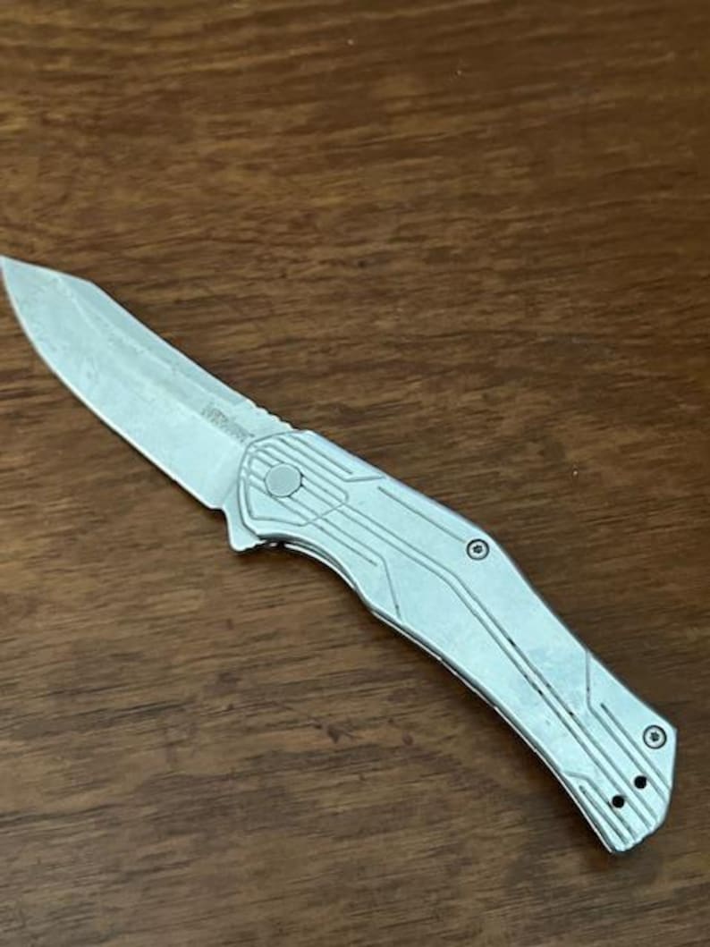 May include: A silver folding knife with a textured blade and handle. The knife has a partially serrated edge and a clip for carrying. The handle features a geometric design and a button for opening and closing the blade.