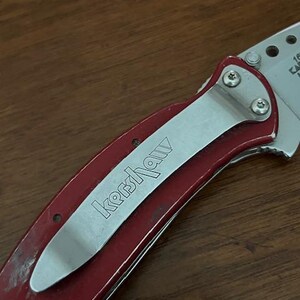 May include: A folding pocket knife with a metallic red handle and a silver pocket clip featuring the engraved Kershaw brand name. The knife has a partially visible silver blade with circular cutouts and small screws securing the clip.