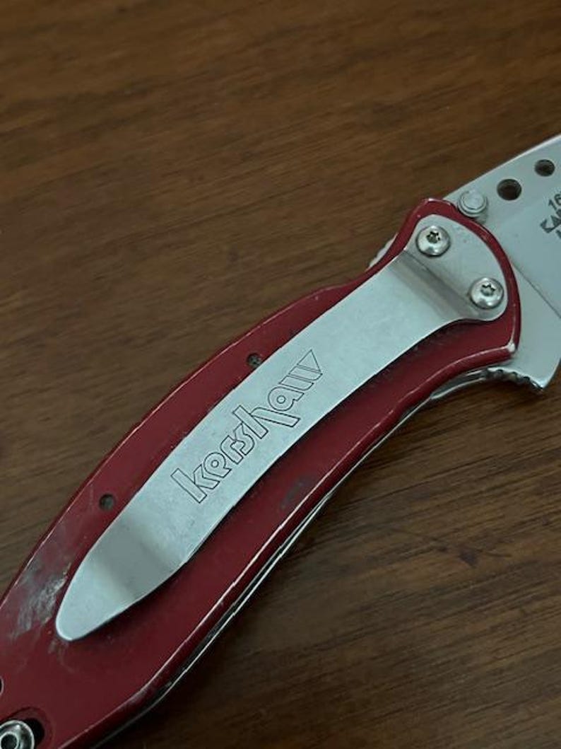 May include: A folding pocket knife with a metallic red handle and a silver pocket clip featuring the engraved Kershaw brand name. The knife has a partially visible silver blade with circular cutouts and small screws securing the clip.