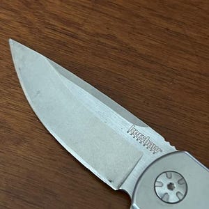 Op de afbeelding: Een zilverkleurig vouwmesblad met een puntige top en een scherpe snede. Het metalen oppervlak is voorzien van de gegraveerde tekst Kershaw SpeedSafe bij het draaipunt, dat een ronde schroefkop met een zespuntig sterpatroon bevat.
