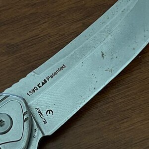 May include: Close-up of a partially visible silver folding knife with a textured blade. The blade has the text "1380" and "Patented" printed on it. The knife is on a wooden surface.