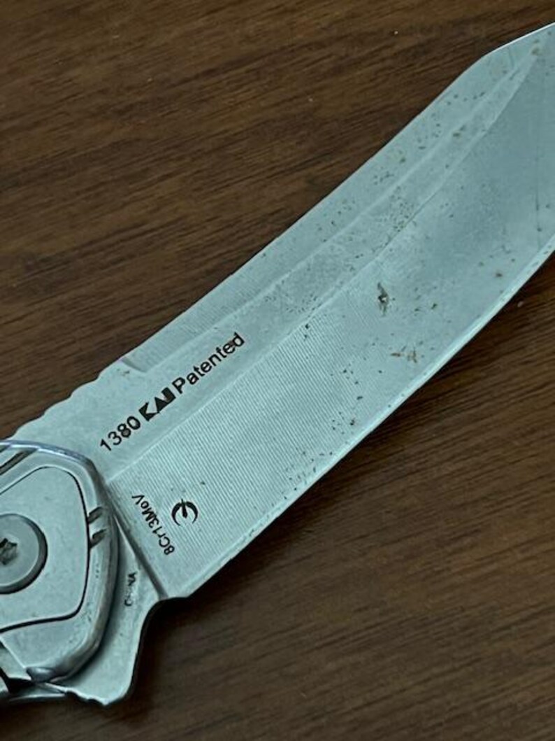 May include: Close-up of a partially visible silver folding knife with a textured blade. The blade has the text "1380" and "Patented" printed on it. The knife is on a wooden surface.
