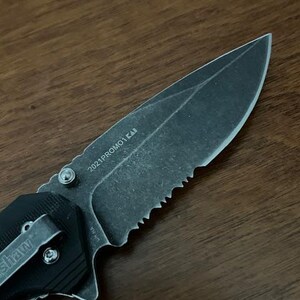May include: A black folding knife with a partially serrated blade. The blade has the text "2021PROMO1 KAB" etched on it. The handle features a pocket clip with the brand name "Kershaw" visible. The knife is set against a dark wood grain background.