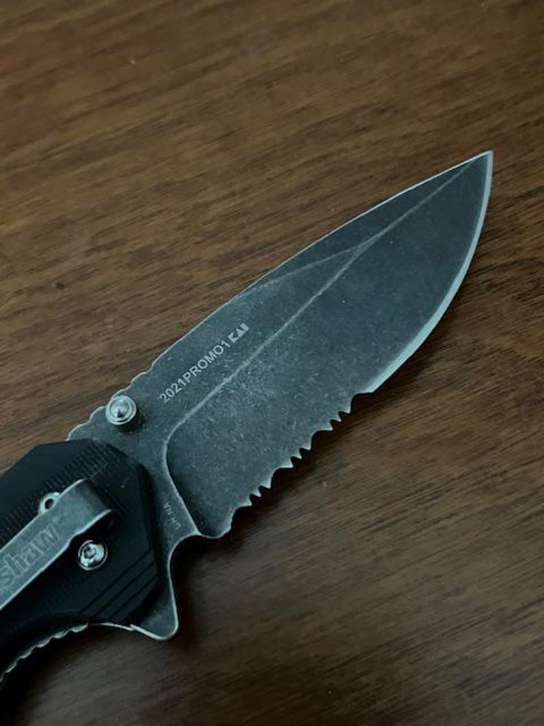 May include: A black folding knife with a partially serrated blade. The blade has the text "2021PROMO1 KAB" etched on it. The handle features a pocket clip with the brand name "Kershaw" visible. The knife is set against a dark wood grain background.
