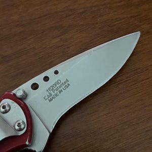 May include: A folding knife with a silver-toned metal blade featuring the text 1620RD, KAI, Patented, and MADE IN USA. The blade has three circular cutouts near the handle, which is accented with a red metallic finish and secured by silver screws.