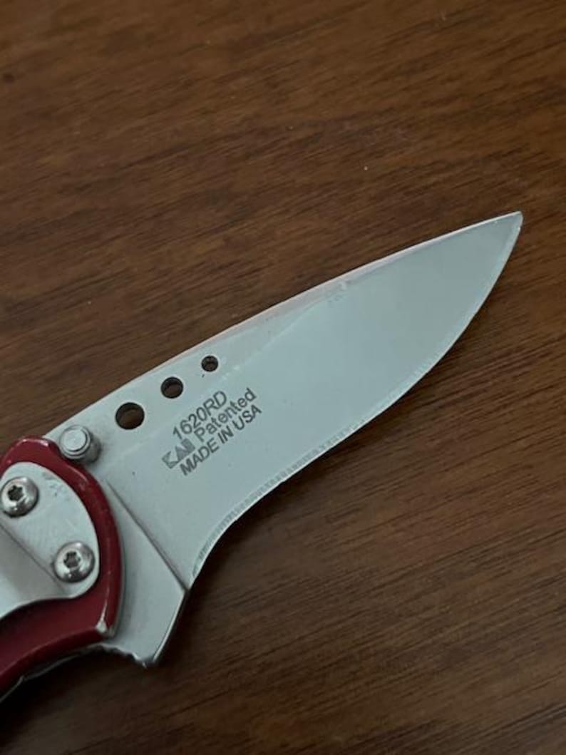 May include: A folding knife with a silver-toned metal blade featuring the text 1620RD, KAI, Patented, and MADE IN USA. The blade has three circular cutouts near the handle, which is accented with a red metallic finish and secured by silver screws.