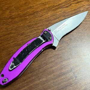 May include: A folding pocket knife with a silver blade and a bright purple handle. The handle has a black clip and is secured with silver rivets. The blade has text that reads "KAB Patented Ken Onion Design".