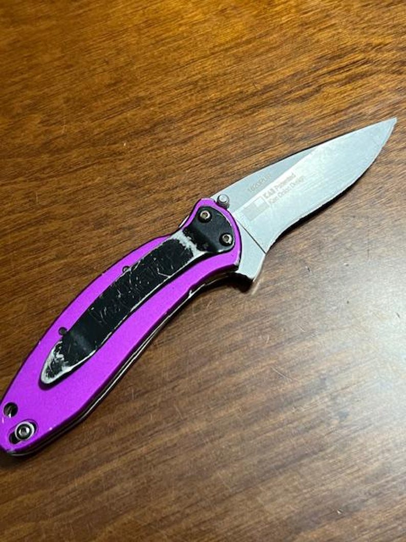 May include: A folding pocket knife with a silver blade and a bright purple handle. The handle has a black clip and is secured with silver rivets. The blade has text that reads "KAB Patented Ken Onion Design".