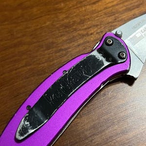 May include: Close-up of a purple folding knife with a black pocket clip. The knife blade is silver and partially visible. The pocket clip has the brand name "Kershaw" printed on it.