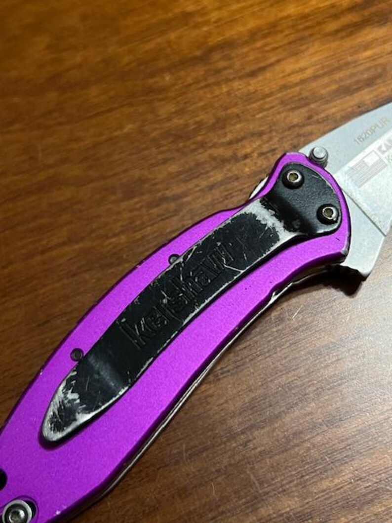 May include: Close-up of a purple folding knife with a black pocket clip. The knife blade is silver and partially visible. The pocket clip has the brand name "Kershaw" printed on it.
