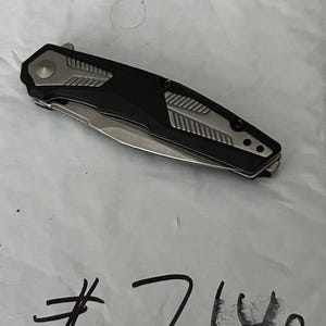 May include: A black and silver folding knife with a partially exposed blade. The handle has a black center with silver accents and a silver button. The number "7140" is handwritten in black ink below the knife.