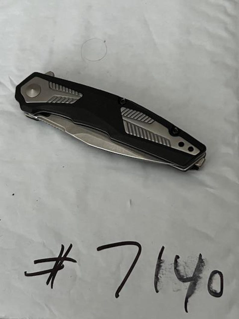 May include: A black and silver folding knife with a partially exposed blade. The handle has a black center with silver accents and a silver button. The number "7140" is handwritten in black ink below the knife.