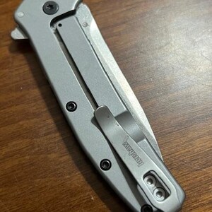 May include: A silver folding pocket knife with a clip. The knife has a blade and a handle with black screws. The clip is marked with the brand name "Kershaw".