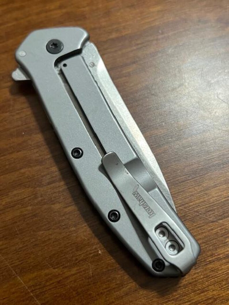 May include: A silver folding pocket knife with a clip. The knife has a blade and a handle with black screws. The clip is marked with the brand name "Kershaw".