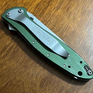 May include: A green folding pocket knife with a silver pocket clip. The word "Kershaw" is printed on the clip. The knife is closed and resting on a wooden surface.