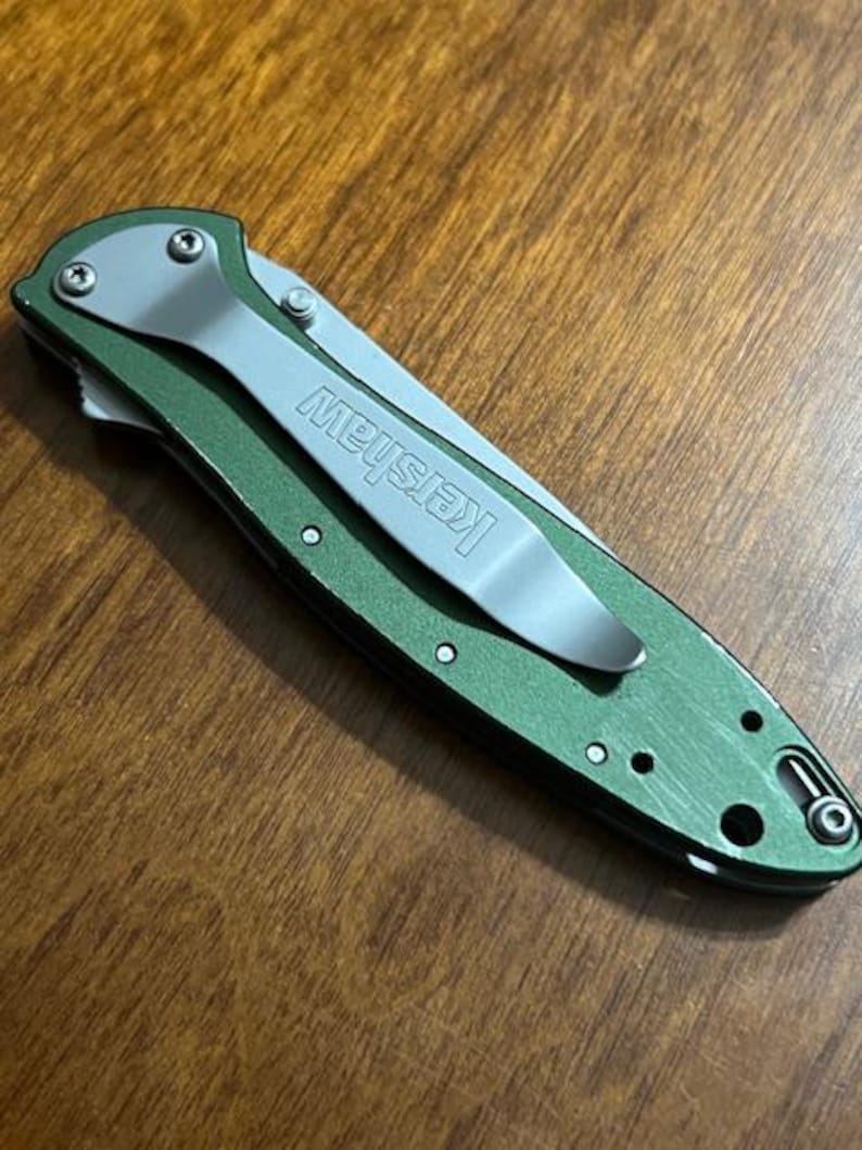 May include: A green folding pocket knife with a silver pocket clip. The word "Kershaw" is printed on the clip. The knife is closed and resting on a wooden surface.