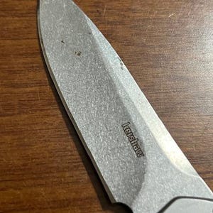 May include: Close-up of a silver Kershaw folding knife blade. The blade is slightly worn and shows the brand name "Kershaw". The knife is set against a brown wooden surface.