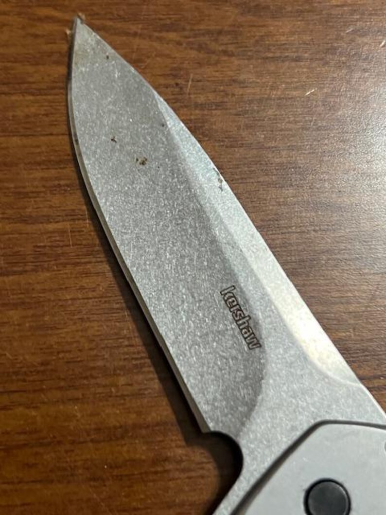 May include: Close-up of a silver Kershaw folding knife blade. The blade is slightly worn and shows the brand name "Kershaw". The knife is set against a brown wooden surface.