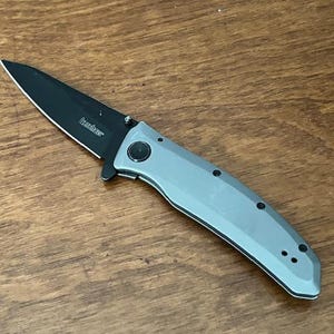 K&ouml;nnte beinhalten: Ein Klappmesser mit schwarzer Klinge und grauem Griff. Auf der Klinge ist der Markenname "Kershaw" aufgedruckt. Das Messer ist ge&ouml;ffnet und liegt auf einer Holzoberfl&auml;che.
