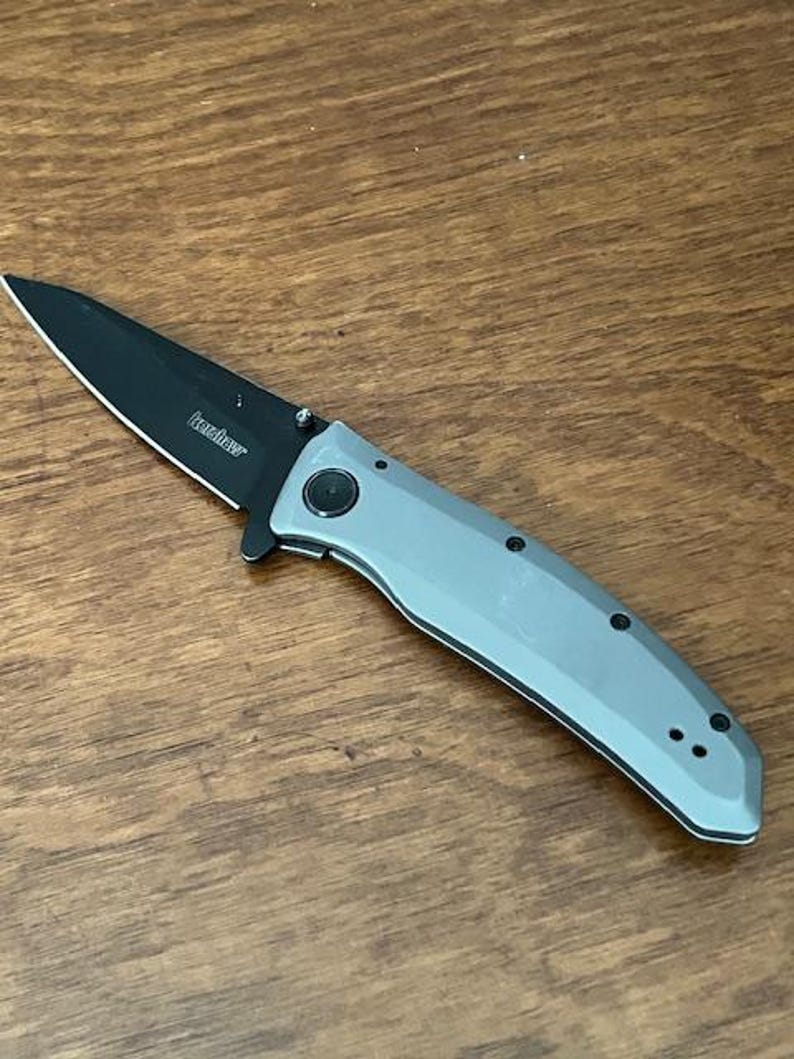 K&ouml;nnte beinhalten: Ein Klappmesser mit schwarzer Klinge und grauem Griff. Auf der Klinge ist der Markenname "Kershaw" aufgedruckt. Das Messer ist ge&ouml;ffnet und liegt auf einer Holzoberfl&auml;che.