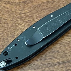May include: Close-up of a black folding knife with a pocket clip. The knife has a partially visible blade and a textured handle. The pocket clip is made of metal and shows signs of wear.
