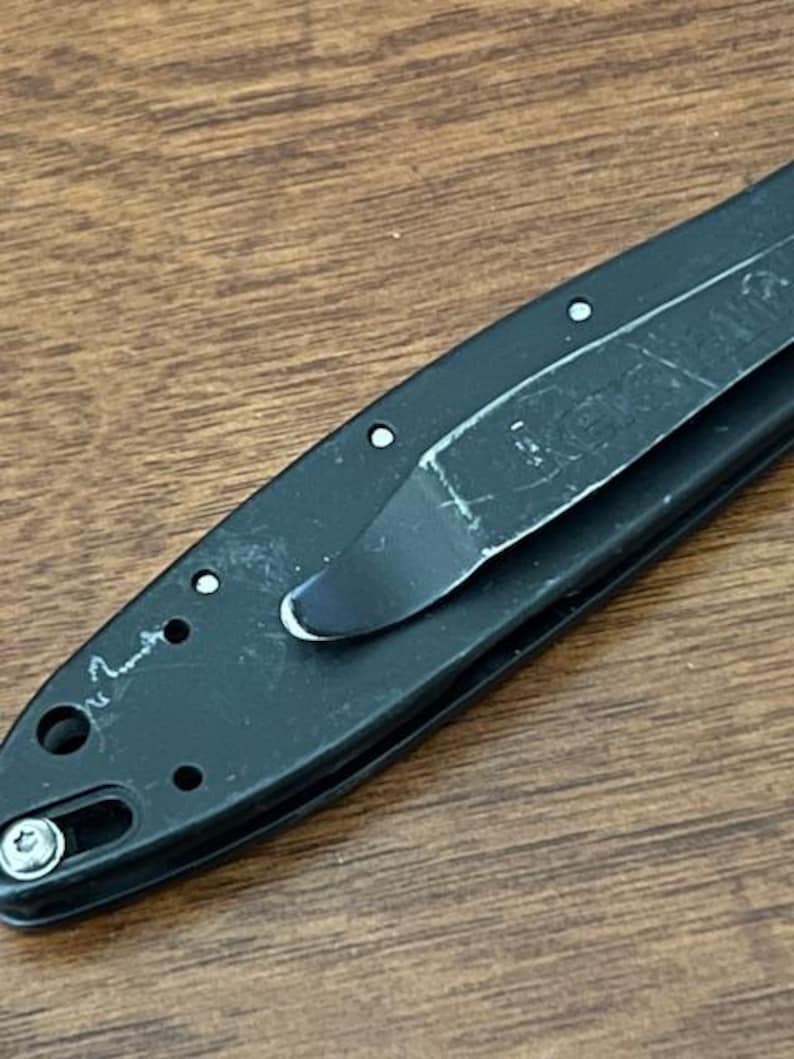 May include: Close-up of a black folding knife with a pocket clip. The knife has a partially visible blade and a textured handle. The pocket clip is made of metal and shows signs of wear.