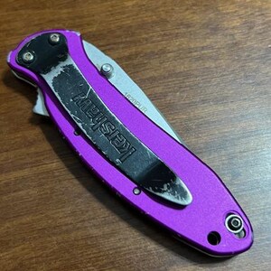May include: A closed purple folding knife with a black pocket clip that reads "Kershaw". The blade is silver and the knife is on a wooden surface. The model number "1620PUR" is visible on the blade.