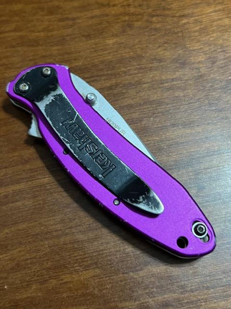 May include: A closed purple folding knife with a black pocket clip that reads "Kershaw". The blade is silver and the knife is on a wooden surface. The model number "1620PUR" is visible on the blade.