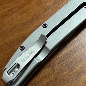 May include: Close-up of a silver folding knife with a pocket clip. The knife has a black accent line and black screws. The pocket clip is engraved with the word "Kershaw". The knife is on a wooden surface.