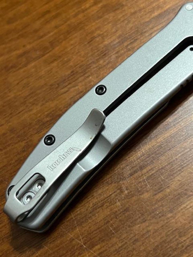 May include: Close-up of a silver folding knife with a pocket clip. The knife has a black accent line and black screws. The pocket clip is engraved with the word "Kershaw". The knife is on a wooden surface.