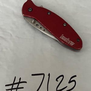 May include: A folding pocket knife with a metallic red handle featuring the Kershaw SpeedSafe USA logo in white text. The silver blade is partially visible, showing a series of small circular cutouts. The number #7125 is written below.