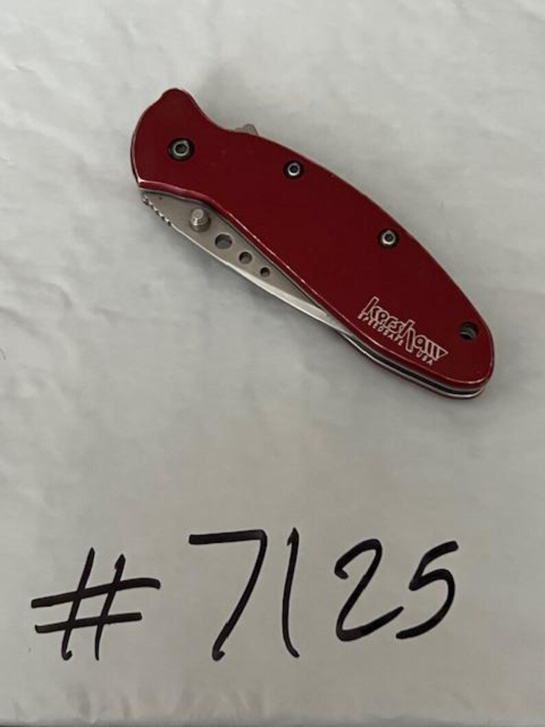 May include: A folding pocket knife with a metallic red handle featuring the Kershaw SpeedSafe USA logo in white text. The silver blade is partially visible, showing a series of small circular cutouts. The number #7125 is written below.