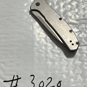 May include: A silver folding knife with a partially exposed blade. The knife has a textured handle with black screws and a small circular button. The number "3020" is written in black marker on the white surface beneath the knife.