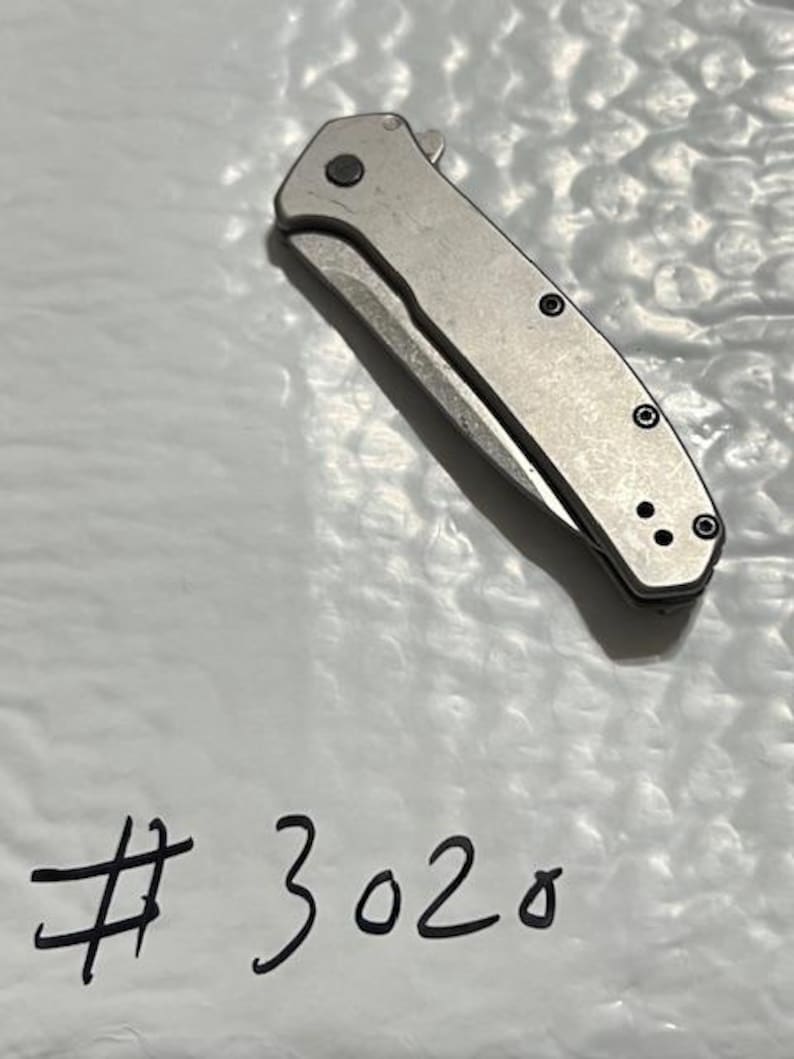 May include: A silver folding knife with a partially exposed blade. The knife has a textured handle with black screws and a small circular button. The number "3020" is written in black marker on the white surface beneath the knife.