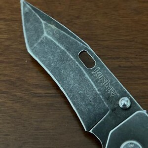 Puede incluir: Una hoja de navaja plegable con un acabado oscuro lavado a la piedra y una punta tanto distintiva. La hoja presenta un recorte ovalado alargado y la palabra Kershaw SpeedSafe grabada en la superficie met&aacute;lica, junto a un tornillo de pivote plateado.