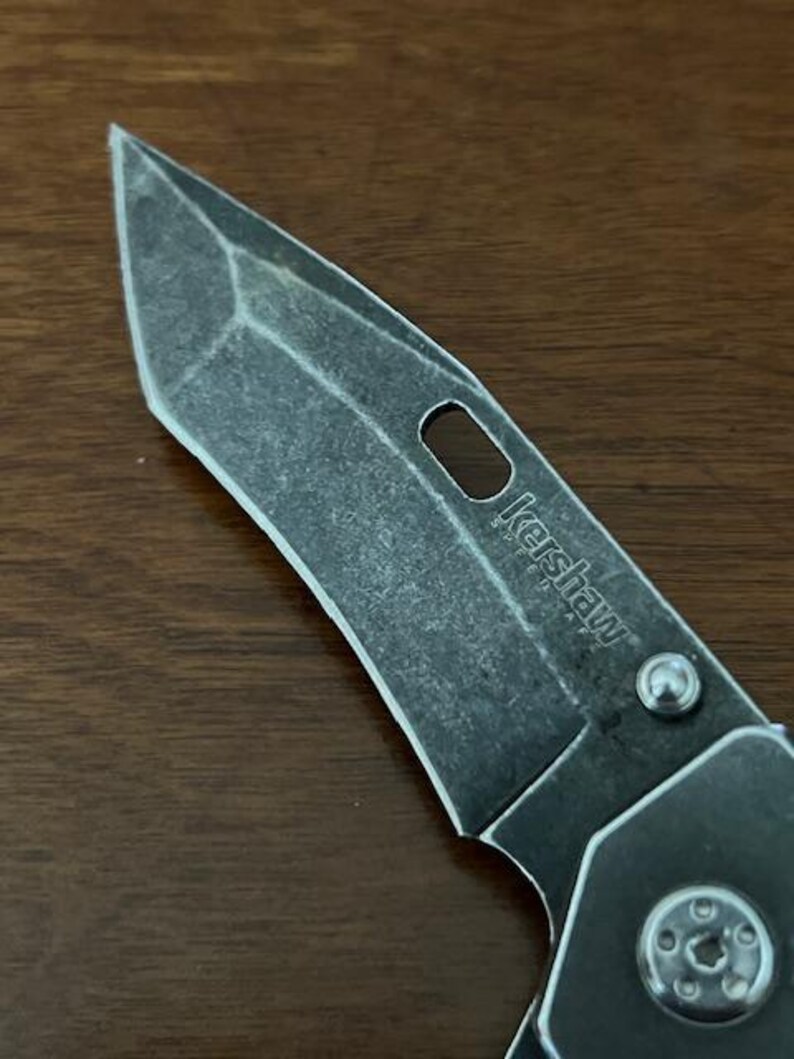 Puede incluir: Una hoja de navaja plegable con un acabado oscuro lavado a la piedra y una punta tanto distintiva. La hoja presenta un recorte ovalado alargado y la palabra Kershaw SpeedSafe grabada en la superficie met&aacute;lica, junto a un tornillo de pivote plateado.