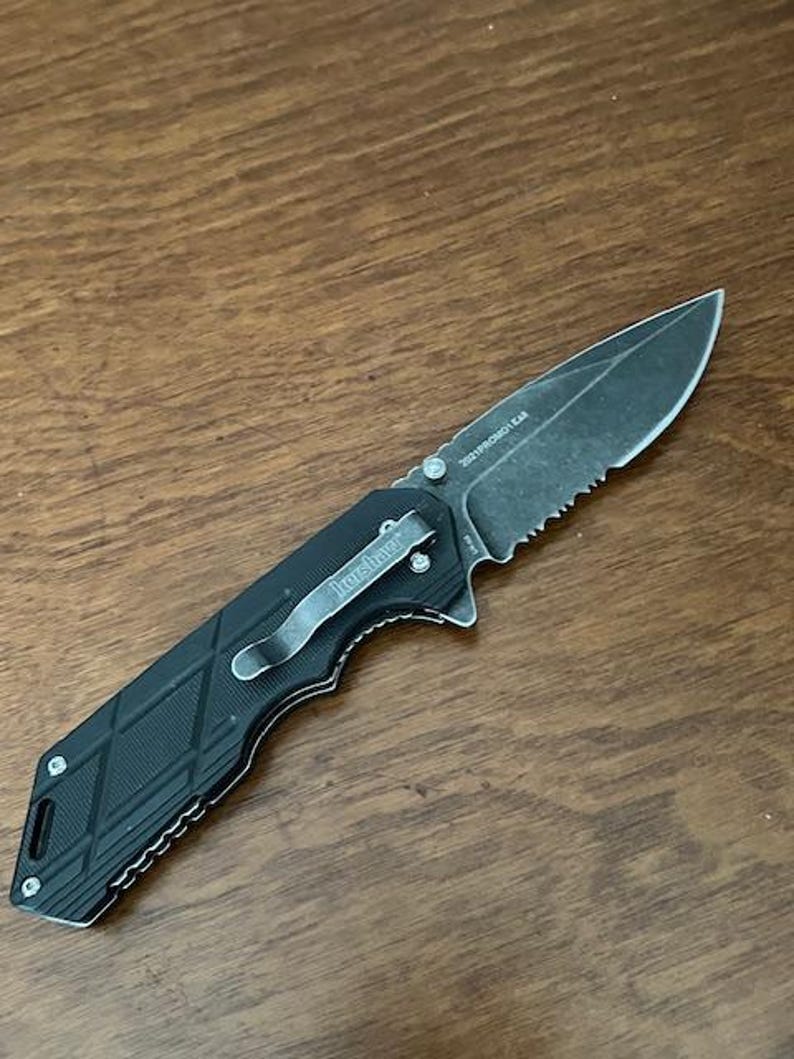 May include: Black folding knife with a partially serrated blade. The knife has a textured black handle with a silver pocket clip that reads "Kershaw". The blade has the text "2021PROMO GEAR".