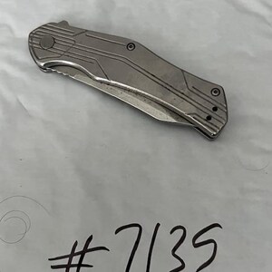 May include: A silver folding knife with a textured handle and a partially serrated blade. The knife has a series of parallel lines etched into the handle. The number "7135" is written in black ink below the knife.