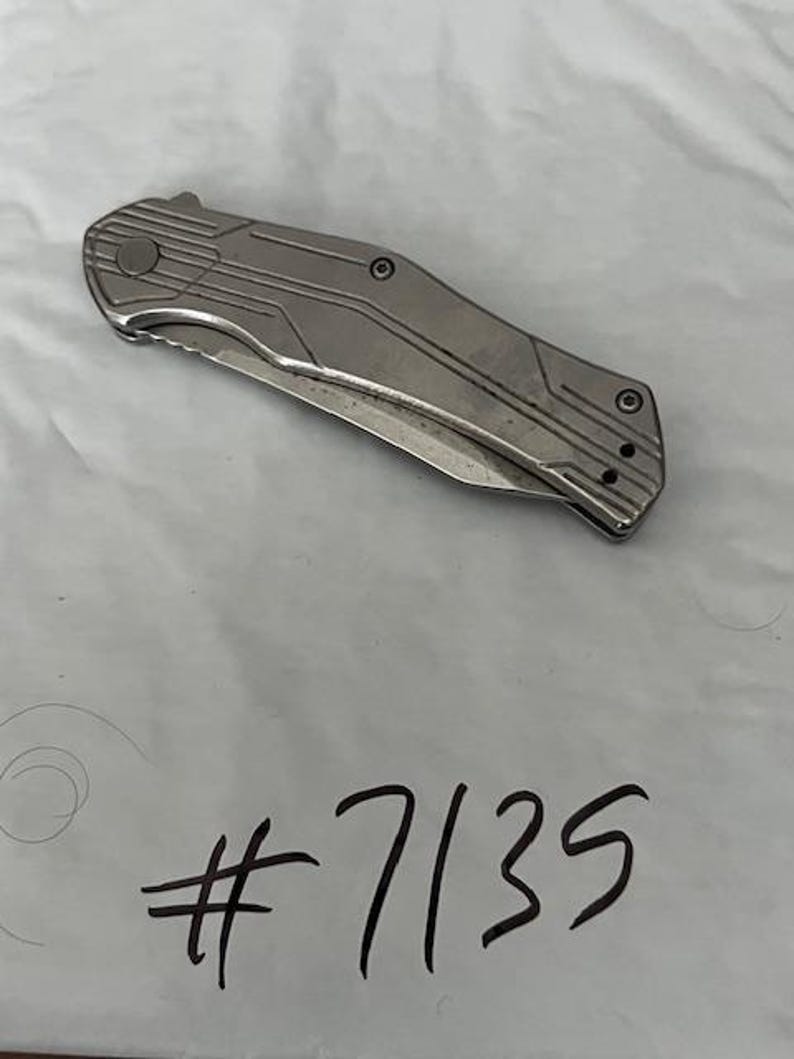 May include: A silver folding knife with a textured handle and a partially serrated blade. The knife has a series of parallel lines etched into the handle. The number "7135" is written in black ink below the knife.
