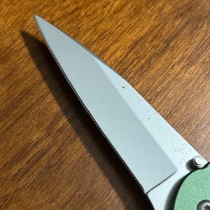 May include: Close-up of a partially visible folding knife. The blade is silver and angled, with a sharp point. The handle is green and has a circular design. The knife is set against a wooden surface.