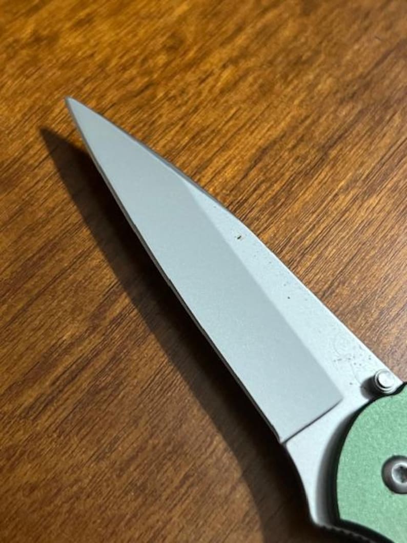 May include: Close-up of a partially visible folding knife. The blade is silver and angled, with a sharp point. The handle is green and has a circular design. The knife is set against a wooden surface.