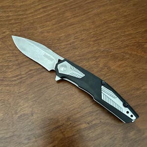 May include: A folding pocket knife with a silver blade and a black and silver handle. The knife is open and angled, displaying the blade and handle design. The knife is on a wooden surface.