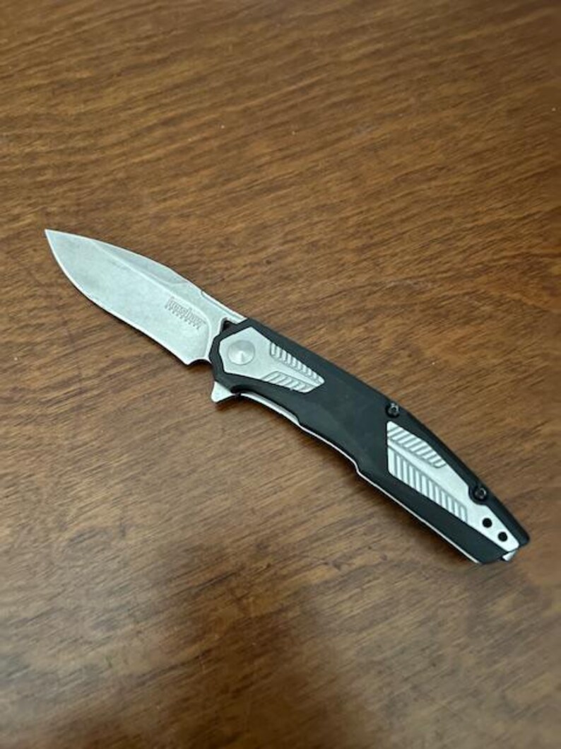 May include: A folding pocket knife with a silver blade and a black and silver handle. The knife is open and angled, displaying the blade and handle design. The knife is on a wooden surface.