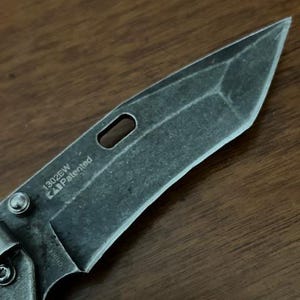 Puede incluir: Una hoja de navaja plegable con un acabado oscuro y desgastado y una punta tipo tanto. La hoja presenta un orificio alargado para el pulgar, un tornillo de pivote y el texto 1302EW, Patented y China grabado en la base del metal.