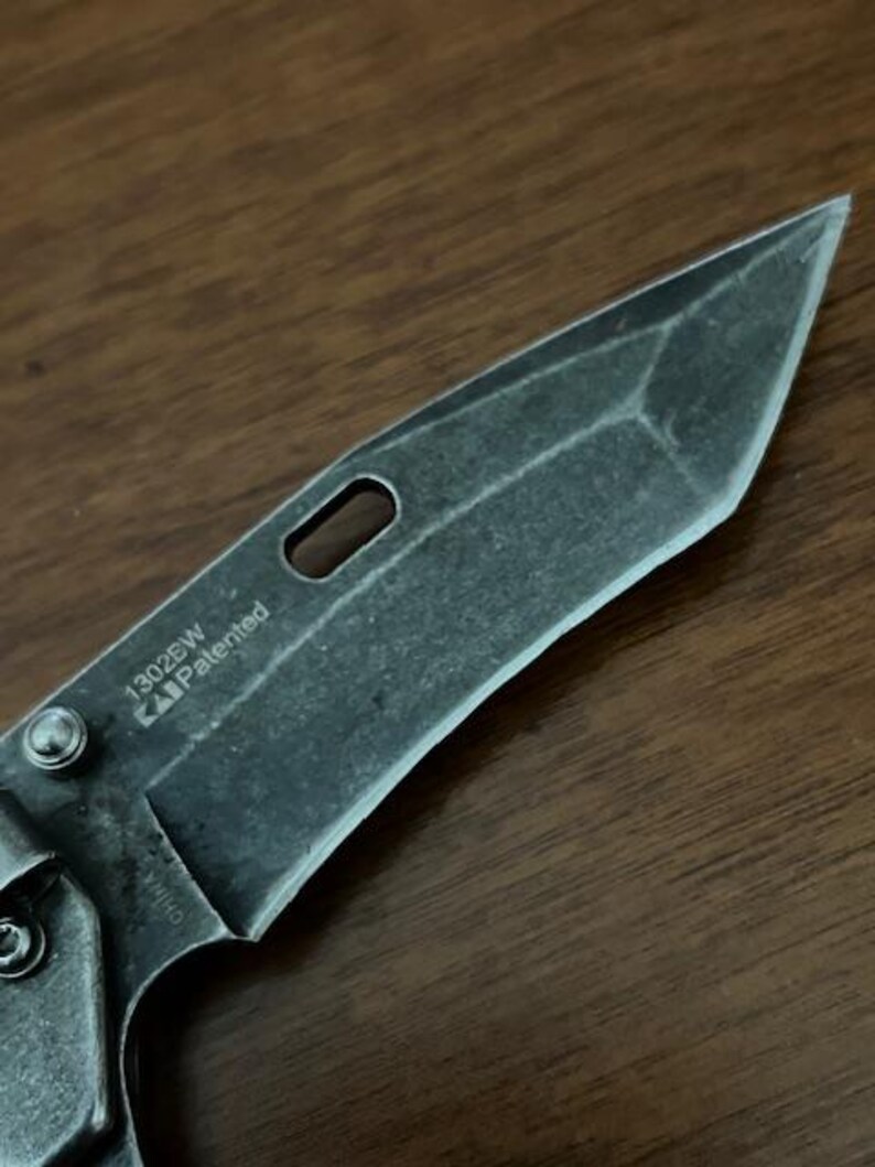 Puede incluir: Una hoja de navaja plegable con un acabado oscuro y desgastado y una punta tipo tanto. La hoja presenta un orificio alargado para el pulgar, un tornillo de pivote y el texto 1302EW, Patented y China grabado en la base del metal.