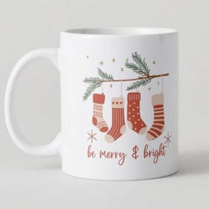 Op de afbeelding: Witte keramische mok met een kerstthema. De mok toont vier hangende kousen in rood en beige, opgehangen aan een groene tak. De woorden "be merry & bright" zijn geschreven in een scriptlettertype.